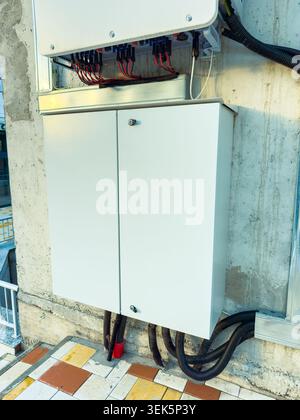 An electrical cabinet is fixed to a wall with multiple wires and cables attached. The scene shows a part of the city during the afternoon. The wall ha Stock Photo