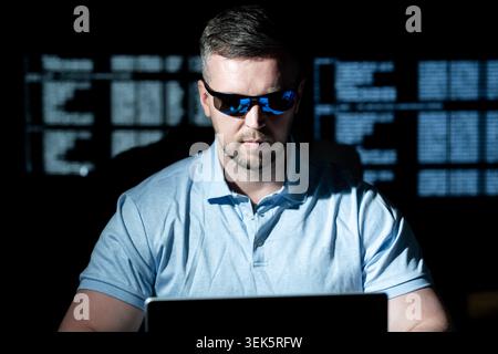 Hacker works on laptop at night. Hacker analyzes code screen in dark room. Hacker monitors network for cyber attack. Hacker targets data system with Stock Photo