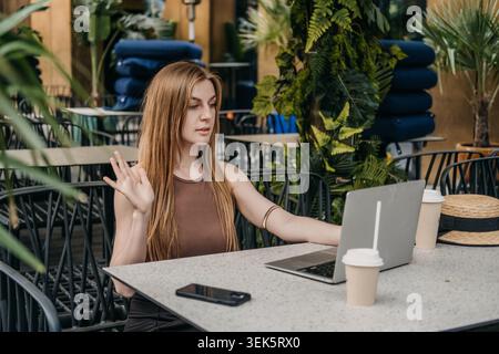 Young woman waves hand during online session on laptop at outdoor cafe. Webinar speaker gesturing, online coaching session, interactive distance Stock Photo