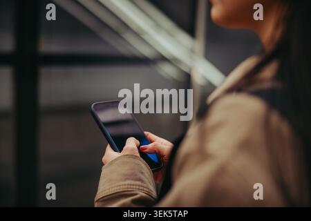 A woman uses her smartphone while waiting on a subway platform, tapping messages and scrolling social media. Hands and screen are in focus, conveying Stock Photo