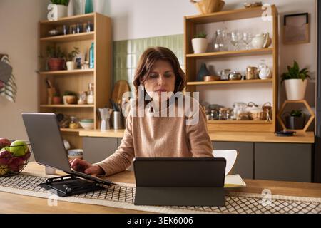 Middle Aged Woman Learning Online at Home Using Laptop and Tablet Stock Photo