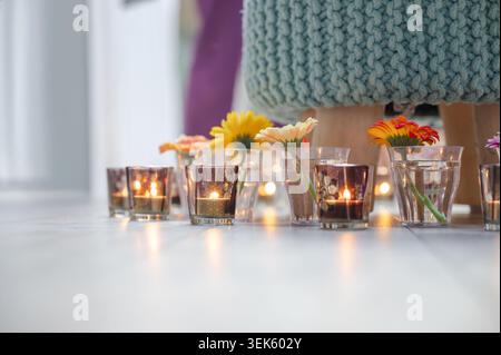 Low angle view of glowing tea light candles in amber glass votives alternating with colorful gerbera flowers on light wooden floor. Turquoise knit pou Stock Photo