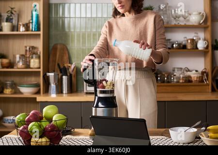 Middle Aged Woman Preparing Smoothie While Learning Online at Home Stock Photo