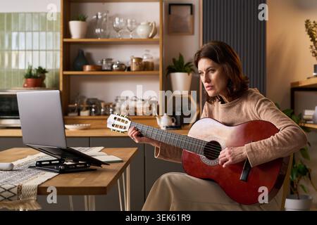 Middle Aged Woman Practicing Guitar during Virtual Lesson at Home Stock Photo