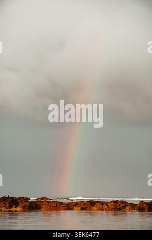 Ujung Genteng Beach features a calm seashore with gentle waves under a dramatic cloudy sky. A subtle rainbow is visible on the horizon Stock Photo