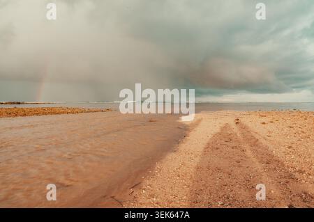 Ujung Genteng Beach features a calm seashore with gentle waves under a dramatic cloudy sky. A subtle rainbow is visible on the horizon Stock Photo