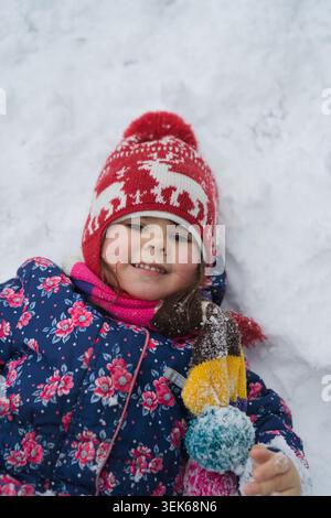 Portrait of smiling girl in red hat lying in snow holding mitten Stock Photo