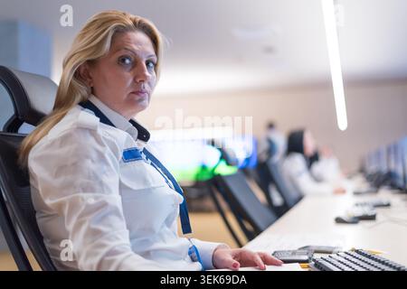 A focused woman in a white security uniform sits at a desk equipped with multiple computer monitors showing CCTV feeds. She is working in a modern, br Stock Photo