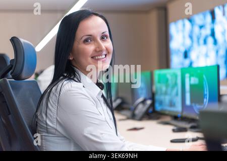 Close-up portrait of a cheerful female security agent smiling while monitoring multiple computer screens in a high-tech surveillance and dispatch cent Stock Photo