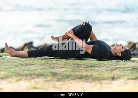 Woman practicing a half wind relieving yoga pose on a mat by the ocean Stock Photo
