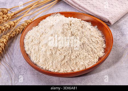 Pile of white flour in rustic wooden bowl. Composition features golden wheat stalks, light pink napkin, and textured grey background. Soft lighting en Stock Photo