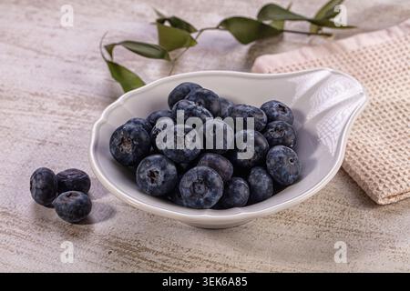 Pile of fresh organic blueberries in elegant white bowl, few berries scattered on light textured wooden surface. Natural light illuminates rich blue p Stock Photo