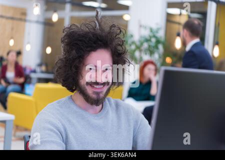 Close-up portrait of a cheerful man with curly hair and a beard, wearing a grey sweater, smiling at the camera while working at a computer in a bustli Stock Photo