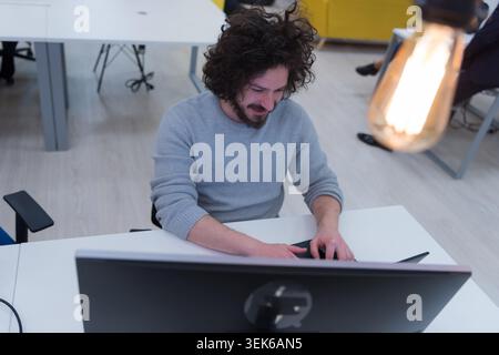 Close-up portrait of a cheerful man with curly hair and a beard, wearing a grey sweater, smiling at the camera while working at a computer in a bustli Stock Photo