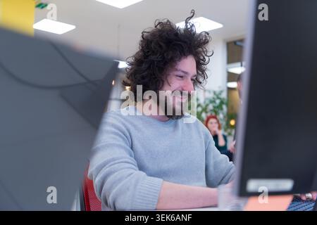 Close-up portrait of a cheerful man with curly hair and a beard, wearing a grey sweater, smiling at the camera while working at a computer in a bustli Stock Photo