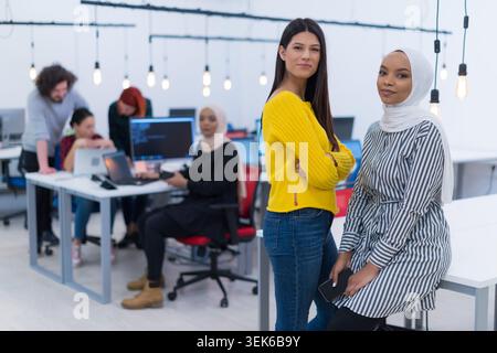 A woman in a yellow sweater and a woman in a hijab stand confidently in the foreground of a bright, active startup office. Stock Photo