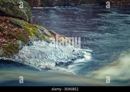 Espoo, Finland - 13.12.2020: Frozen textures on moss-covered boulders beside a rushing river in a wintry landscape Stock Photo