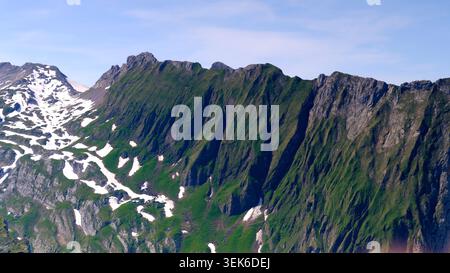 Mountains rise steeply under a blue sky with patches of snow and green grass in this scenic landscape during daytime hours Stock Photo