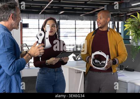 Diverse coworkers discussing VR headset component while holding tablet and full headset at office. Collaboration, prototype, device, hardware, daylight, workspace, technology Stock Photo