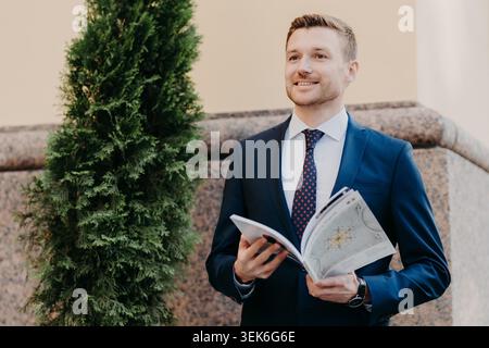 Horizontal shot of attractive male with joyful expression, being focused into distance, has stubble, dressed in formal clothes, holds city map, tries to orientate in big unknown city has smile on face Stock Photo