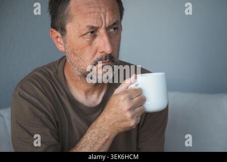 Pensive middle-aged man drinking coffee while sitting indoors with thoughtful, worried expression, selective focus Stock Photo