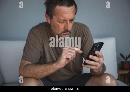 Focused middle-aged man using smartphone on couch at home checking messages and browsing, selective focus Stock Photo