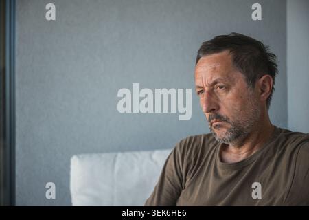 Pensive middle-aged man sitting on couch at home looking thoughtful and worried, contemplative mood, selective focus Stock Photo