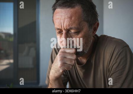 Middle-aged man sits on a sofa with his hand on his chin, appearing pensive and worried. The quiet indoor portrait conveys contemplation, loneliness, Stock Photo