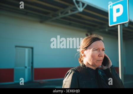 serious woman talking on phone in parking lot near metal canopy outdoors, selective focus Stock Photo
