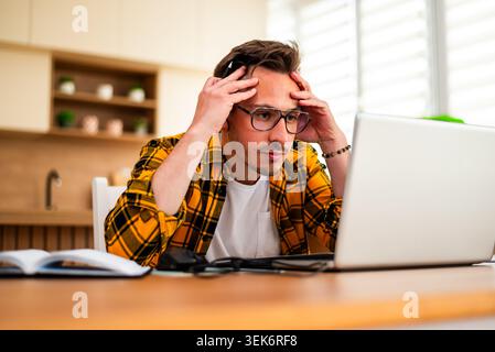 A concerned young man stares at his laptop screen with both hands on his head, appearing overwhelmed by a problem or unexpected information. Stock Photo