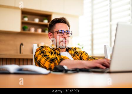 With a pensive expression, a man types on his laptop, appearing to analyze information or solve a problem online. Stock Photo