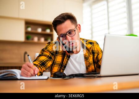 Working from home, a young man pauses to write down ideas and reminders, combining online research with traditional note-taking. Stock Photo