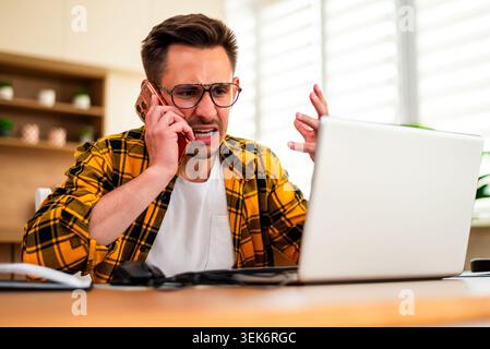 Working from home, a man in a yellow plaid shirt uses his smartphone alongside his laptop, managing multiple tasks in a modern and productive workspac Stock Photo