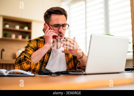 Working from home, a man in a yellow plaid shirt uses his smartphone alongside his laptop, managing multiple tasks in a modern and productive workspac Stock Photo