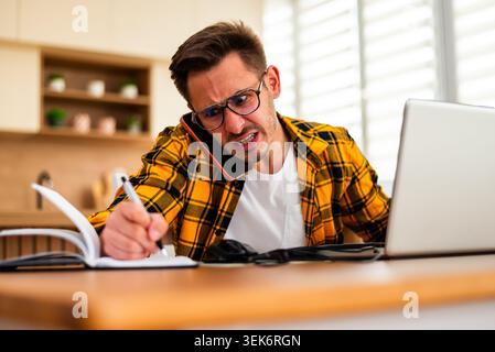 Freelancer is working from home, he is talking on phone and writing notes. His face suggests anger and frustration. Stock Photo