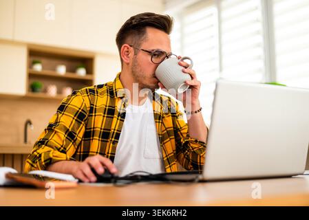 Freelance remote worker is working on his laptop at his home and drinking coffee. He is exhausted. Stock Photo