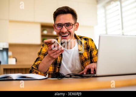 Happy young man is working from his home. He is using laptop and talking on smartphone. Stock Photo