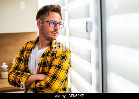 Portrait of young man who is thoughtful. He is looking through window at his home. Stock Photo
