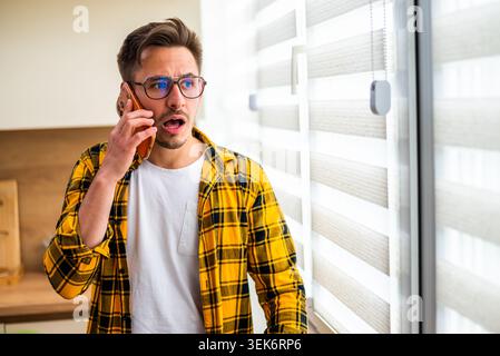 Young man is talking on phone at his home. He appears shocked by the news. Stock Photo