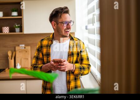 Standing near a window, a cheerful young man in a yellow plaid shirt typing message on his smartphone. Stock Photo