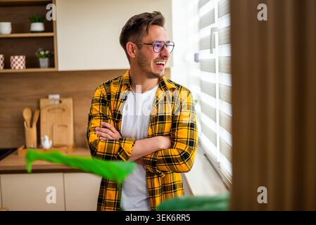 Portrait of happy young man with arms crossed. He is looking through window in his kitchen. Stock Photo