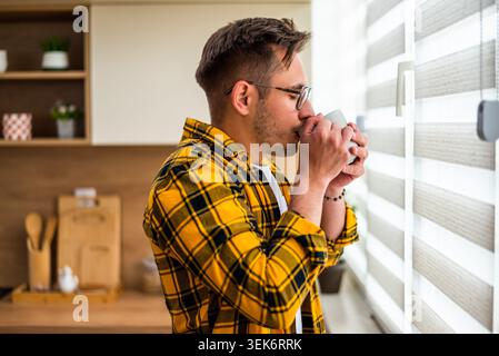 Young man is standing by the window in his kitchen and drinking coffee. Stock Photo
