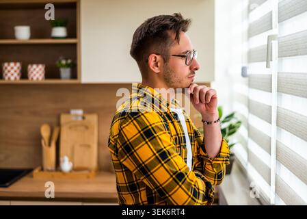 A concerned man pauses his daily routine to gaze out the window, appearing overwhelmed by a challenge or personal matter. Stock Photo