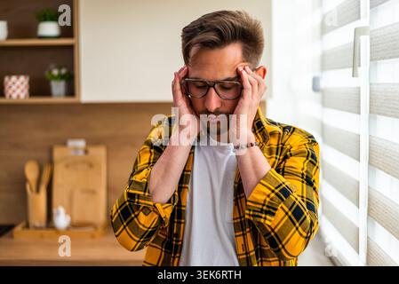 A stressed young man stands by the window with both hands on his head. The image conveys anxiety, uncertainty, and concern about an important situatio Stock Photo