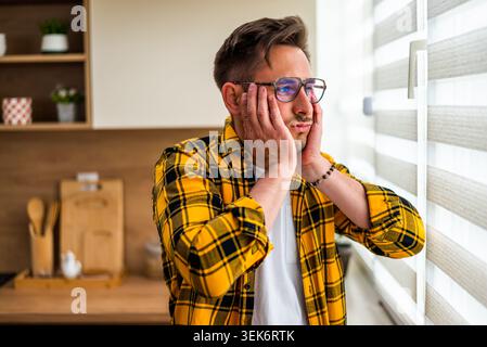 A stressed young man stands by the window with both hands on his cheeks, looking outside with a worried expression. The image conveys anxiety, uncerta Stock Photo