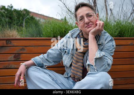 A woman sits on a wooden bench, relaxed and smiling, surrounded by grass. Stock Photo