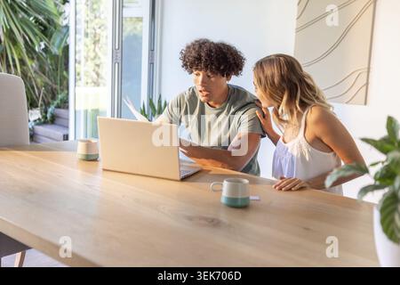 Diverse couple reviewing documents and using laptop at wooden dining table with ceramic mugs Stock Photo