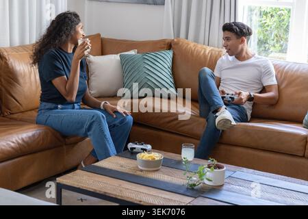 African American couple casually dressed sitting on leather sofa at home holding controller Stock Photo