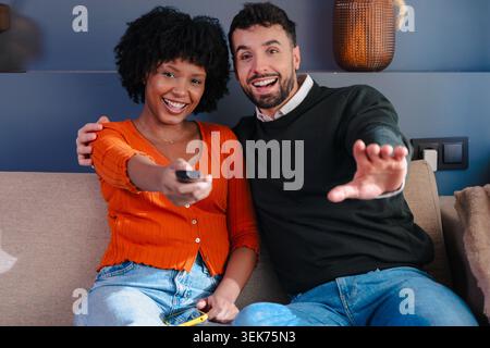 Happy multiracial couple watching television and using remote control on sofa Stock Photo