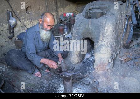 Blacksmith sitting, hammering hot metal on an anvil near a traditional forge Stock Photo